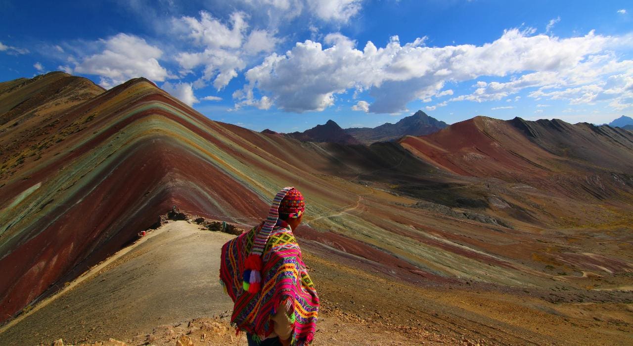 A person dressed in traditional Peruvian clothing stands with their back to the camera in front of Rainbow Mountain or Vinicunca in Peru